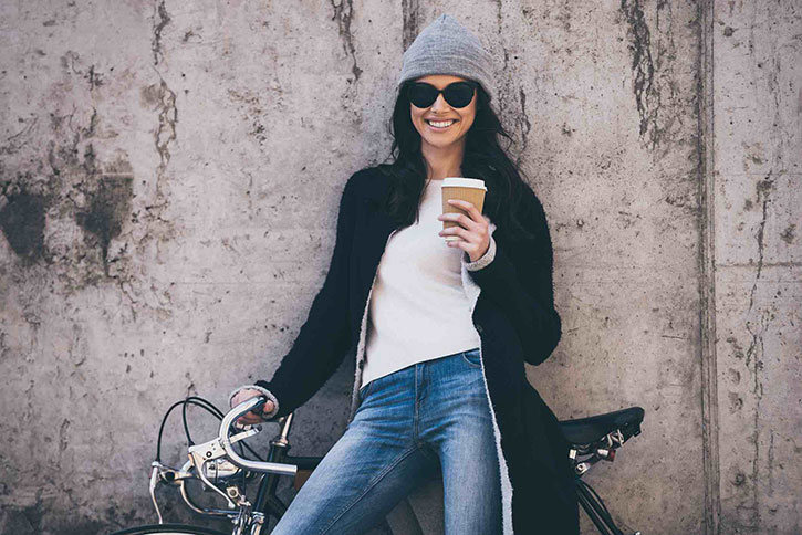 Eine gut gelaunte Frau lehnt mit Ihrem Fahrrad gegen einer Wand. In der Hand hält sie einen Becher Kaffee Eine gut gelaunte Frau lehnt mit Ihrem Fahrrad gegen einer Wand. In der Hand hält sie einen Becher Kaffee